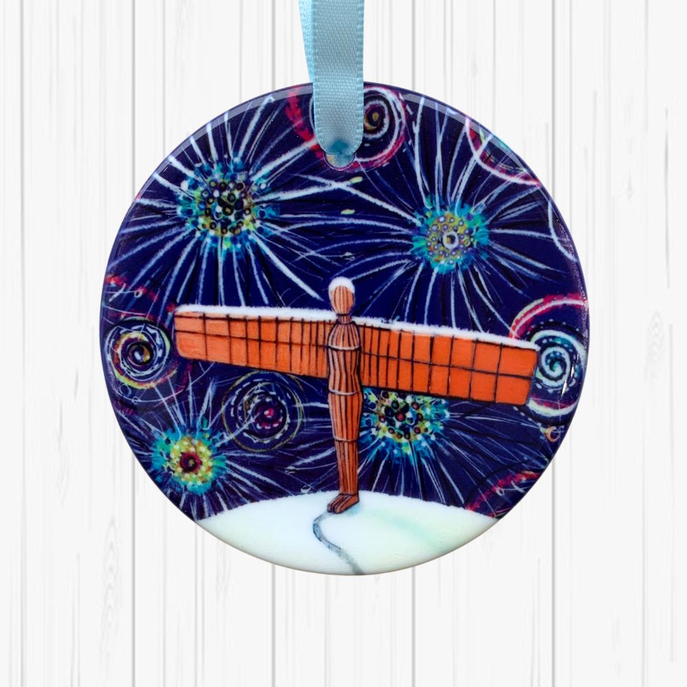 Circular ceramic decoration against a white textured background. It features the Angel of the North on a snowy hill, under a dark sky of colourful fireworks.