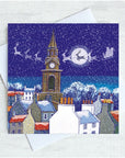 A square Christmas greeting card featuring a snowy night in Berwick village scene with tower, rooftops, sleighs, and Santa in the sky, shown with a white envelope on a light textured background.