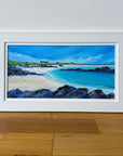 A framed landscape painting of a coastal beach with turquoise water, white sand, dark rocky foreground, and a blue sky, displayed against a white wall on a wooden floor.