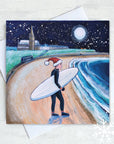 A cheerful surfer approaches the winter waves at Longsands Beach, Tynemouth, as snowflakes fall under a full moon illuminating the sandy beach.