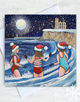 A trio enjoy a festive swim in the waves at King Edward's Bay in Tynemouth. The card features Tynemouth priory in the distance on a cliff and a full moon over the lighthouse. Snow is falling in the dark night sky.