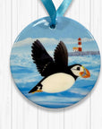 Round ornament with a puffin taking flight and lighthouse design on a blue background.
