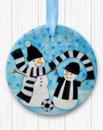 Circular ceramic decoration hung against a white background. Happy snowmen are wearing black and white scarves and football kit with a football at their feet.