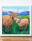 Framed artwork of two highland cows in a field with mountains.