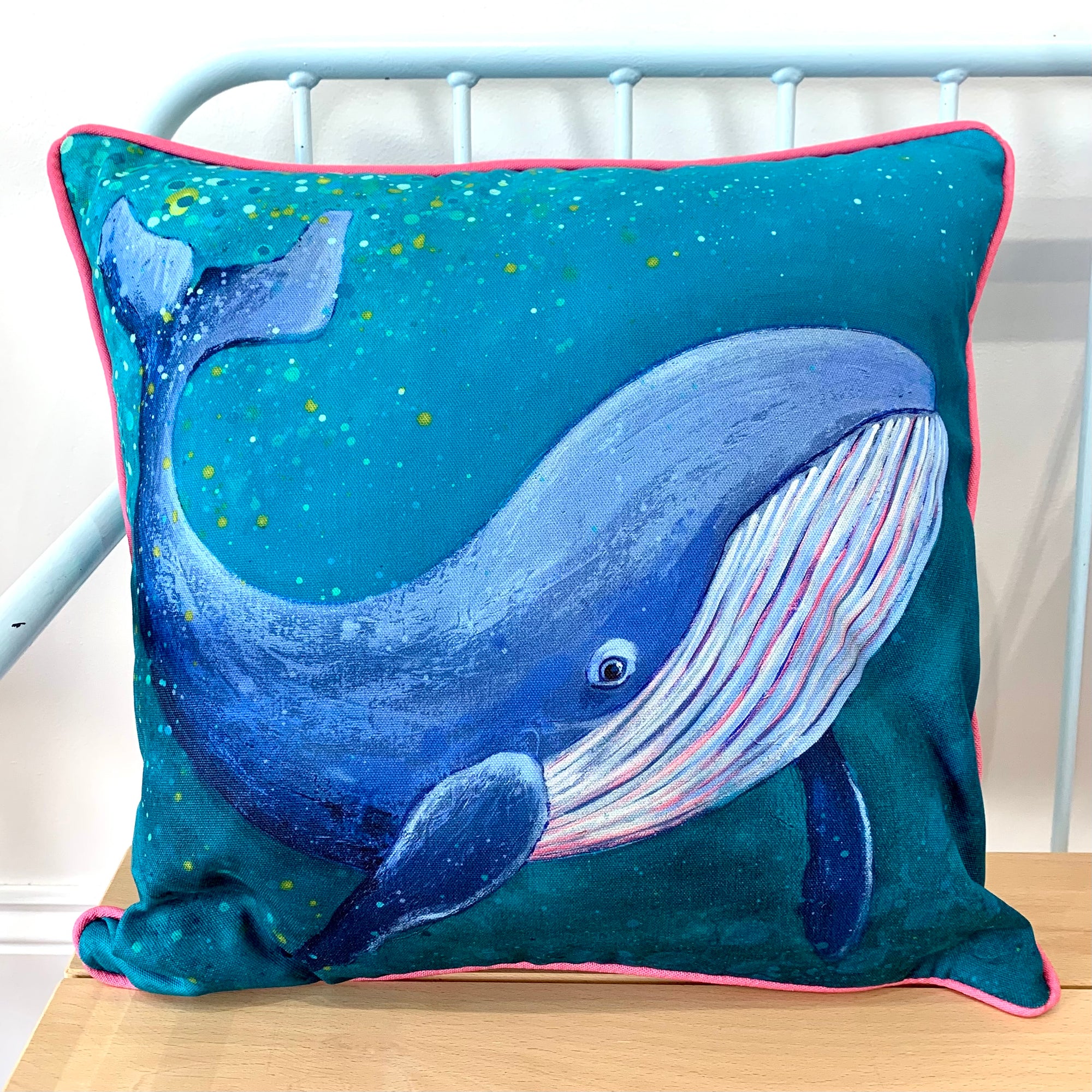 A cushion featuring a blue whale painting on a teal green background with bright pink piping.