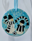Circular ceramic decoration hung on a white branch. Happy snowmen wearing black and white scarves and football kit with a football at their feet.