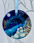 Circular ceramic hanging decoration on a white tree branch. Two smiling snowmen stand side by side by the river with the Tyne Bridge in the background. The sky is dark with a full moon and snow is falling.