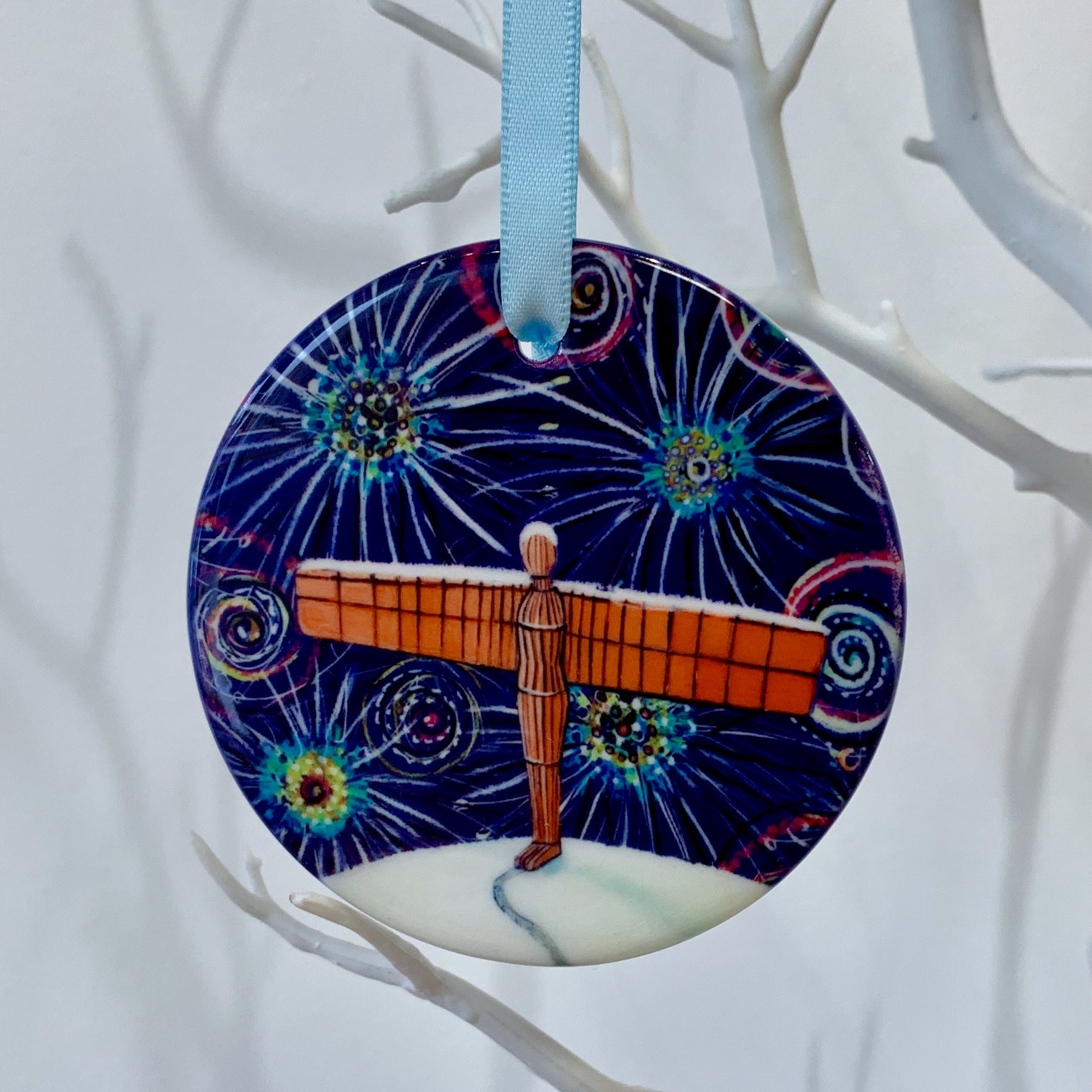 Circular ceramic decoration hung on a white branch. It features the Angel of the North on a snowy hill, under a dark sky of colourful fireworks.