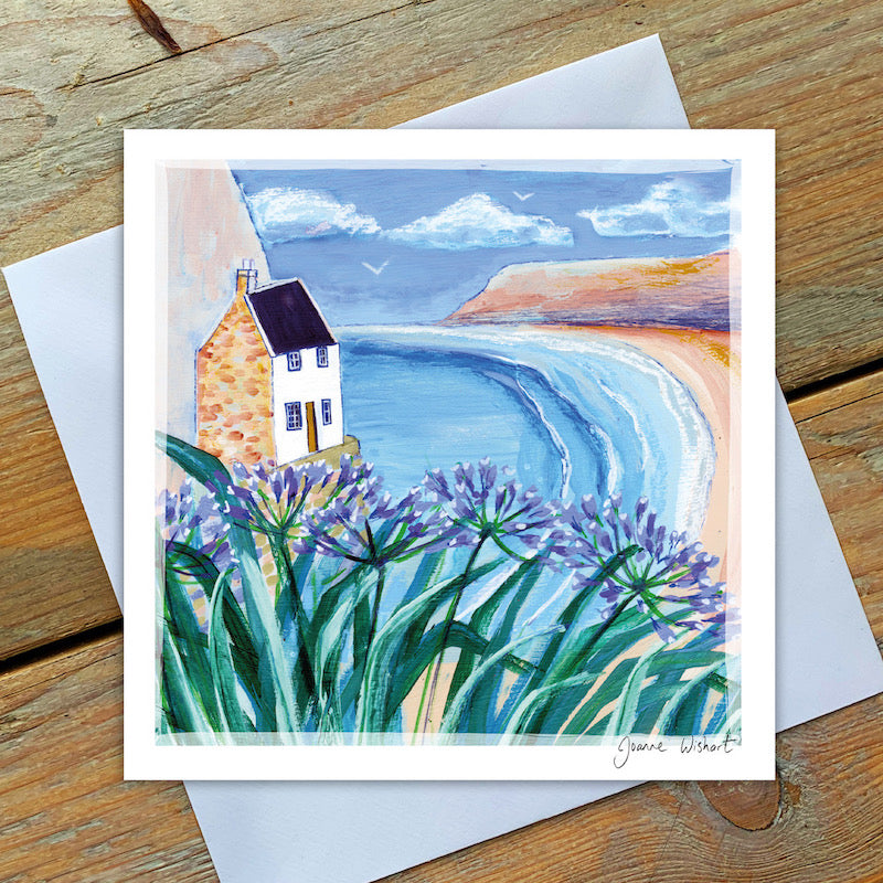 Purple Agapanthus flowers frame a seaside scene of a small whitewashed cottage overlooking a golden beach, calm aquamarine sea and russet cliffs. 