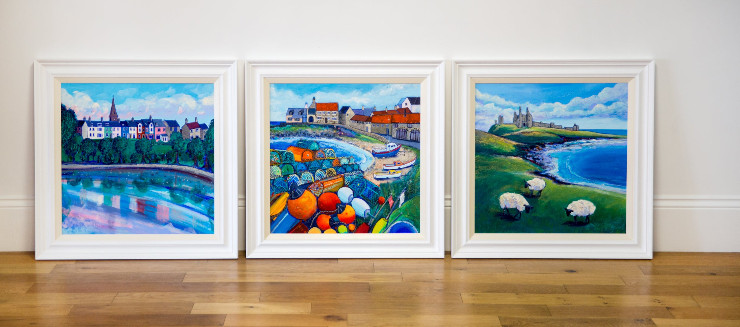 A Row of Three colourful paintings of Northumberland in white wood frames. From left to right Alnmouth featuring a row of colourful cottages. Centre painting is Craster Harbour featuring creels and crails and right the painting features Dunstanburgh Castle with three sheep grazing in the foreground.e