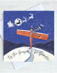 A square Christmas greeting card with a white envelope, featuring an illustration of the Angel of the North and a black-and-white striped scarf standing on a snowy hill under a dark blue night sky with Santa and reindeer flying overhead. The card reads, 'Tis the Season to be Geordie'.