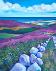 Blanketed hills of purple heather aredived by a stone path which lead down to the valley below. in the distance you can see a colourful patchwork of fields with the bright blue sea beyond. f