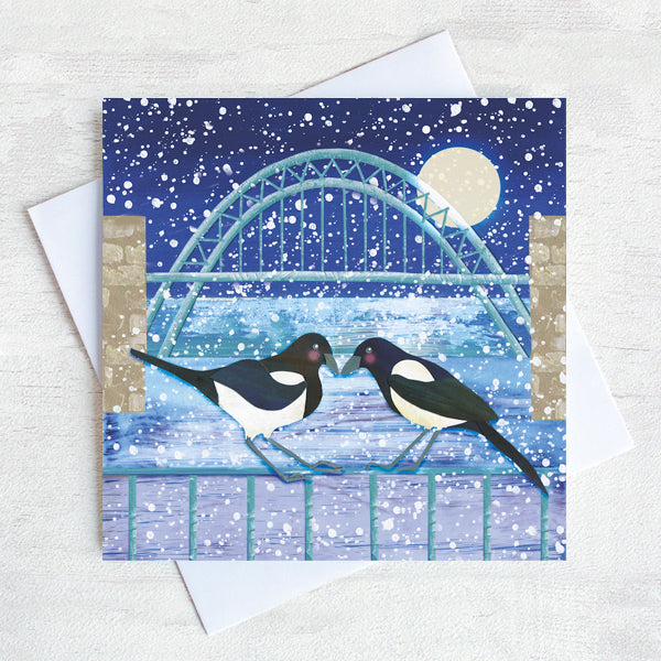 A Christmas card featuring two magpies touching beaks under the Tyne Bridge. snow is falling all around. 