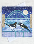 A Christmas card featuring two magpies touching beaks under the Tyne Bridge. snow is falling all around.