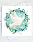 A white dove flies holding and olive branch. It is surrounded by a circular wreath of green foliage.
