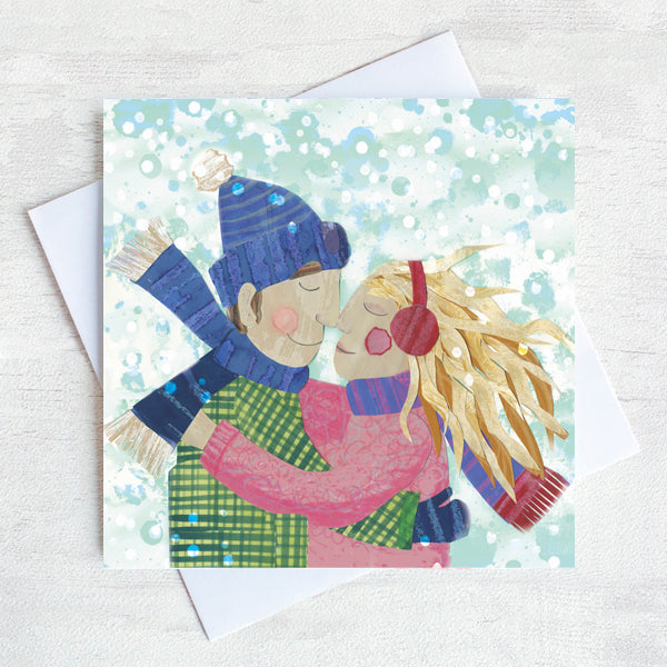A traditional Christmas card featuring a loving couple rubbing noses int he snow. They are wearing snow hats and mit tens and her long yellow hair blows in the snow. s 