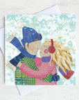 A traditional Christmas card featuring a loving couple rubbing noses int he snow. They are wearing snow hats and mit tens and her long yellow hair blows in the snow. s