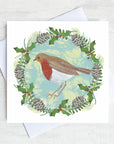A robin with a bright red breast sits in a festive wreath of pine cones, holly and red berries.