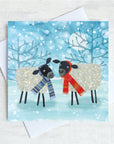 A christmas greetings card featuring two black headed sheep facing one another wearing brightly coloured scarfs. They are standing in a field of show with snowflakes falling all around them.