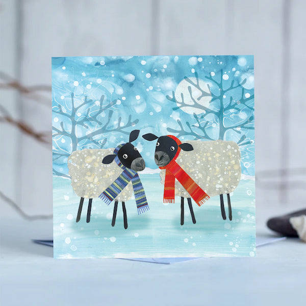 A funny festive card featuring two quirky sheep standing in a snow storm. They are wearing brightly striped scarfs.