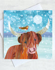 A Christmas card featuring a Highland Cow and a robin perched on one if its horns. THey stand in a fields with snow fluttering overhead.
