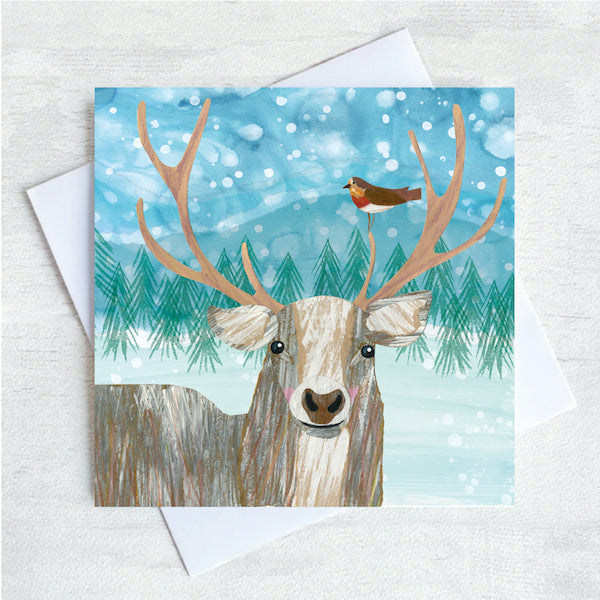 A Christmas Card featuring a stag and a robin perched on it's Antler. A green fir tree forrest is in the background with a flurry of snowfall overhead. 