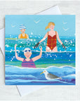 Three ladies and a seagull splash amongst the waves at the seaside. The are wearing broightly coloured swimsuits and have rosy red cheeks.