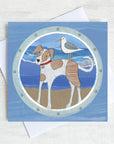 A greetings card featuring a small dog on the beach with a seagull on his back.