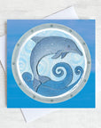 A greetings card featuring a jumping dolphin as seen through a ships porthole.