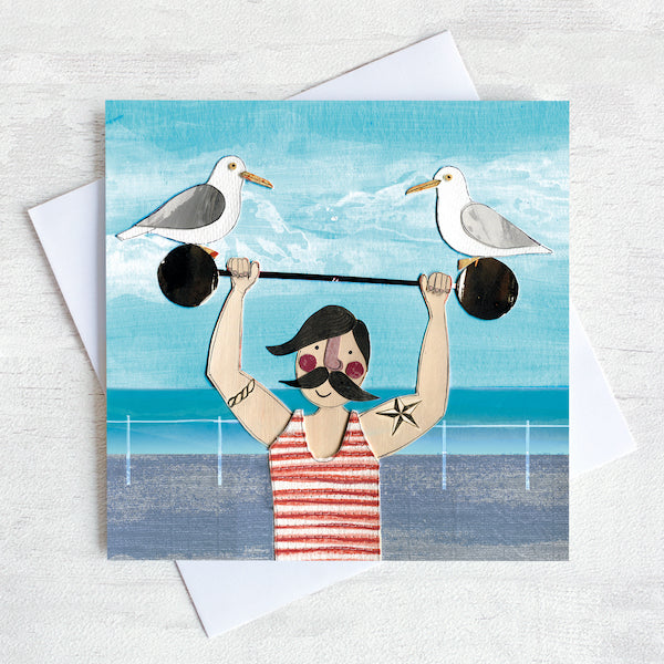 A greetings card of a dapper chap in a stripy vest lifting some weights on the sea front with two cheeky seagulls perched on top.