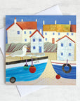 A greetings card featuring white harbour cottages with orange roofs and blue coble boats on the shore with a cheeky seagull perched.