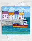 A charming coastal greetings card featuring colourful cottages on a harbour wall with a fishing boat bobbing in the water.