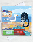 A coastal greetings card with a cute cat and seagull sitting on a harbour wall looking to eat the catch of the day