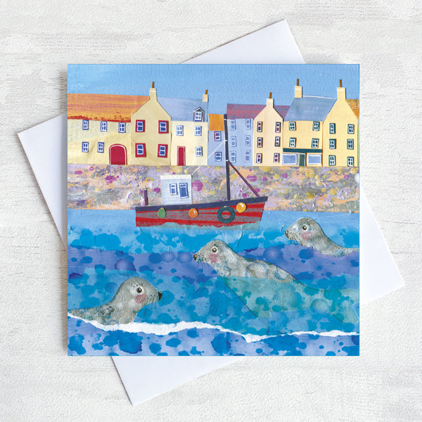 A greetings card of coastal harbour featuring seals bobbing in the water around a fishing boat.