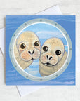 A greetings card featuring two cheeky seals peeking through a ships porthole.
