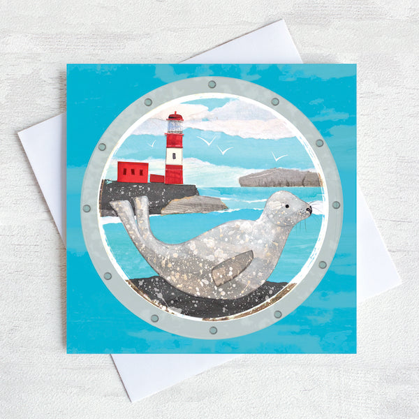 A greetings card featuring a grey seal basking on the rocks in front of a red and white lighthouse.