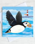 A coastal greetings card with a flying puffin swooping over a quirky red and white lighthouse.