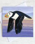 A quirky greetings card featuring a flying puffin gliding over the sea in the evening light.