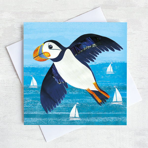 A flying puffin with its wings spread over the turquoise sea on an illustrated greetings card.