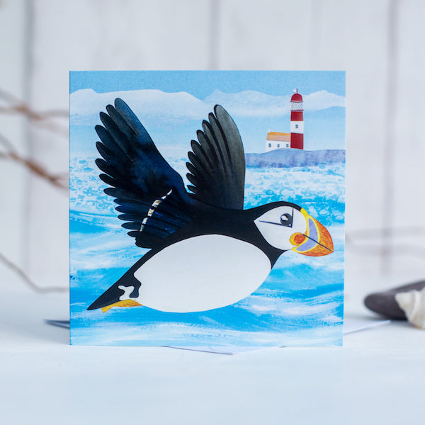 A greetings card featuring a flying puffin in front of a quirky little lighthouse.