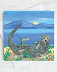 A greetings card with a sea otter curled on a moonlit beach surrounded by seaweed.