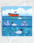 A seaside greetings card with a seals head bobbing in the sea.