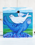 A greetings card featuring a cute family of seals snuggled on each others back, with a black headed gull on top.
