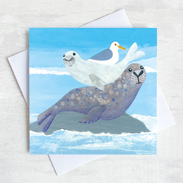 A seal basking on a pack with it's pup and a cheeky sea gull on it's back. This is an illustrated card by Joanne Wishart.