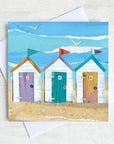 A colourful coastal greetings card featuring 3 beach huts all in a row on a golden sand beach.