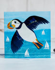 A greetings card showing c colourful puffin flying over the turquoise sea. White sailing boats bob on the water below.