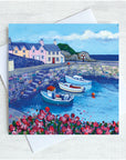 Three fishing boats sit in the harbour with some pasetel coloured houses above the harbour wall. Pink flowers adorn the foreground.