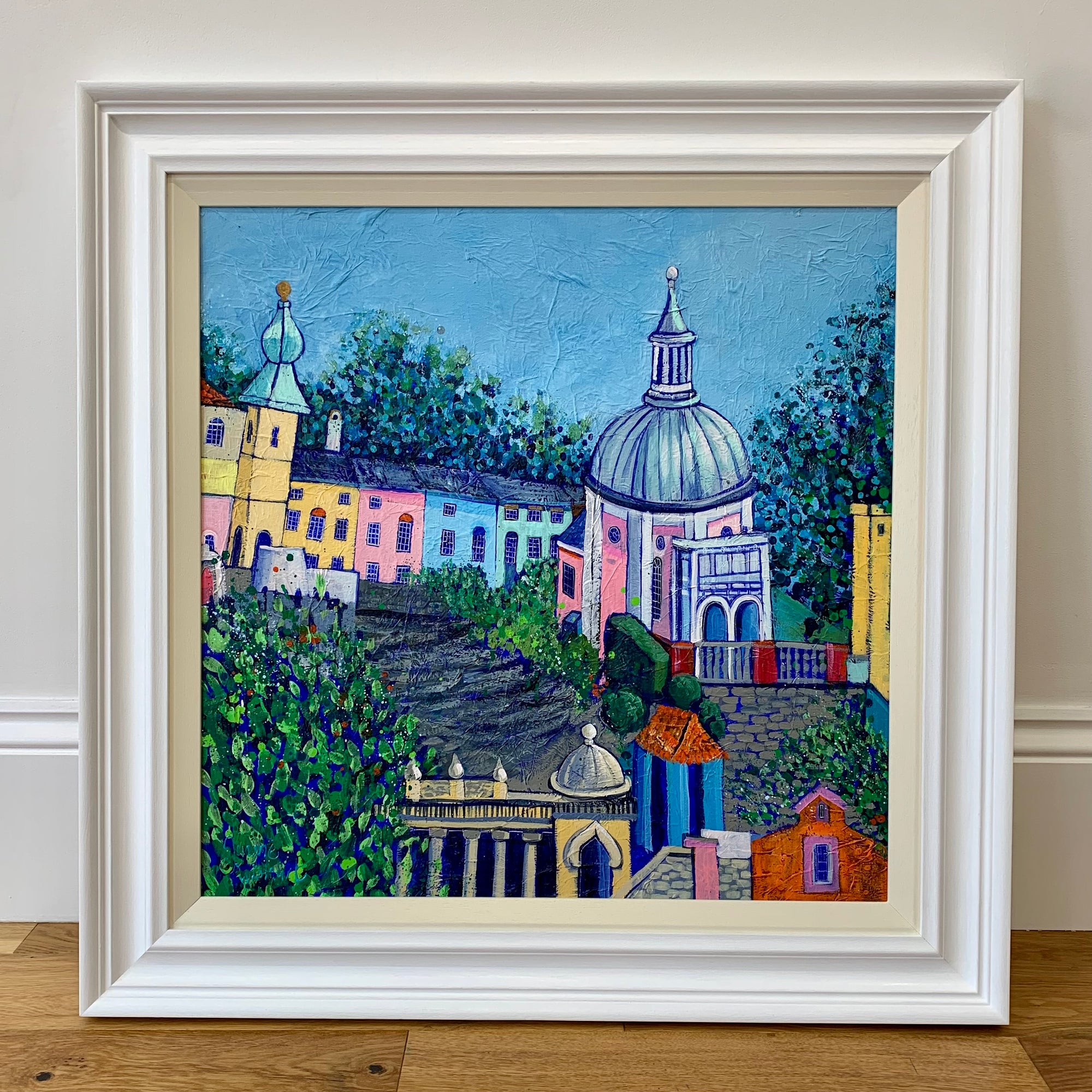 A painting in a white wood frame featuring the colourful italian village of Portmeirion.  