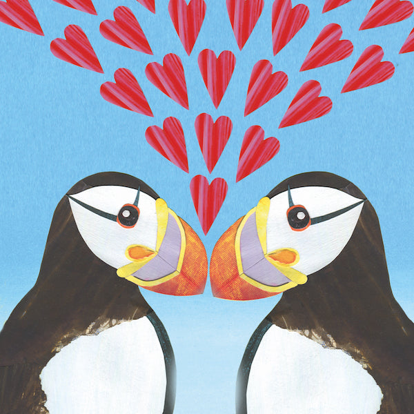 Two puffins stand beak to beak with multiple textured red love hearts between them. The puffins have their characteristic markings of bright orange and yellow beaks with white chests and faces. 