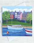 a white sail boat sits on the water infornt of a row of brightly coloured houses. THe grass is green and the tress are in full bloom.